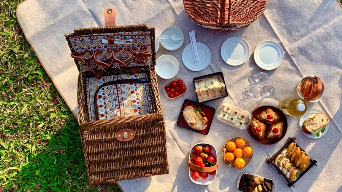 An open picnic basket with lots of picnic foods on a blanket on a sunny afternoon