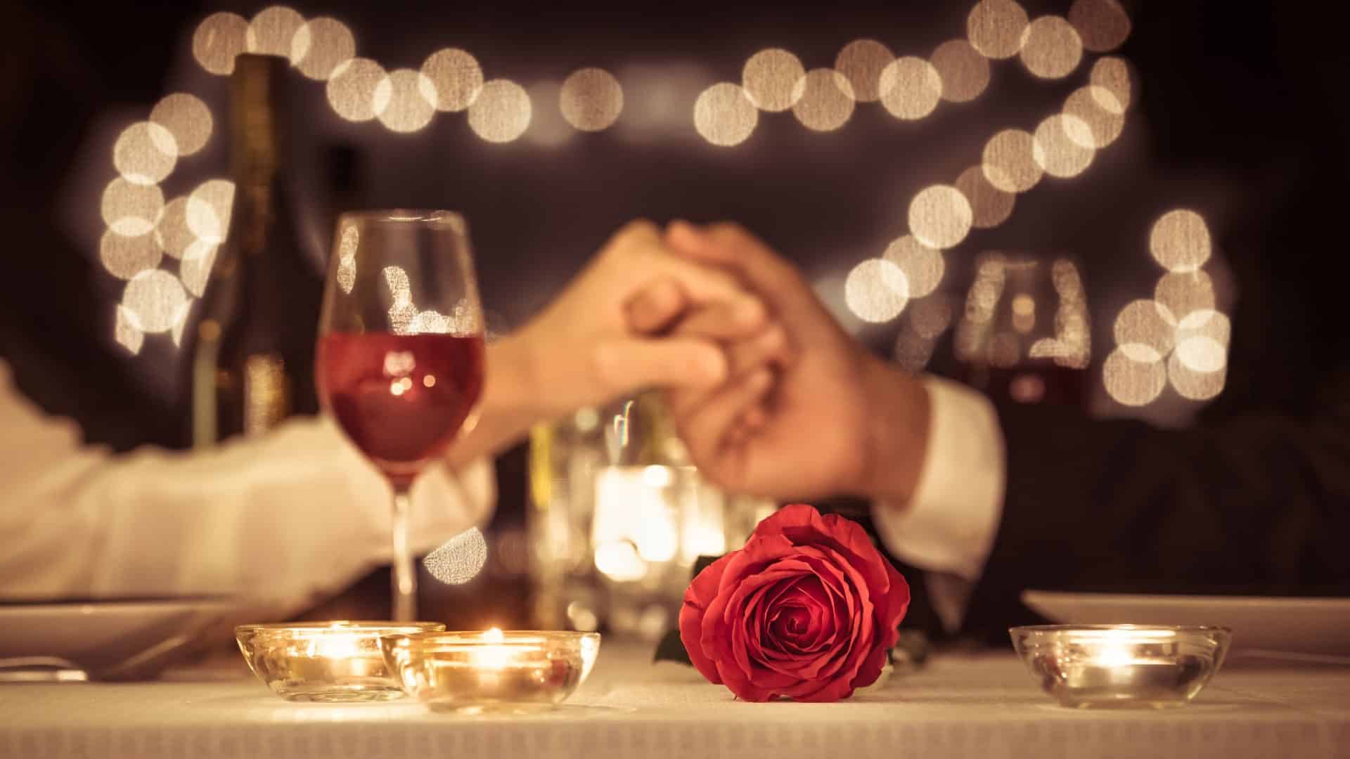 Couple holding hands across a romantic, candlelit dinner table with red wine and a red rose.
