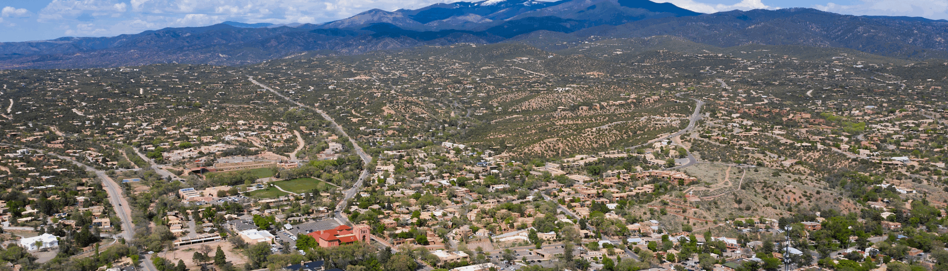 view of santa fe new mexico, with a mix of adobe-style buildings desert and surrounding mountains under a partly cloudy sky