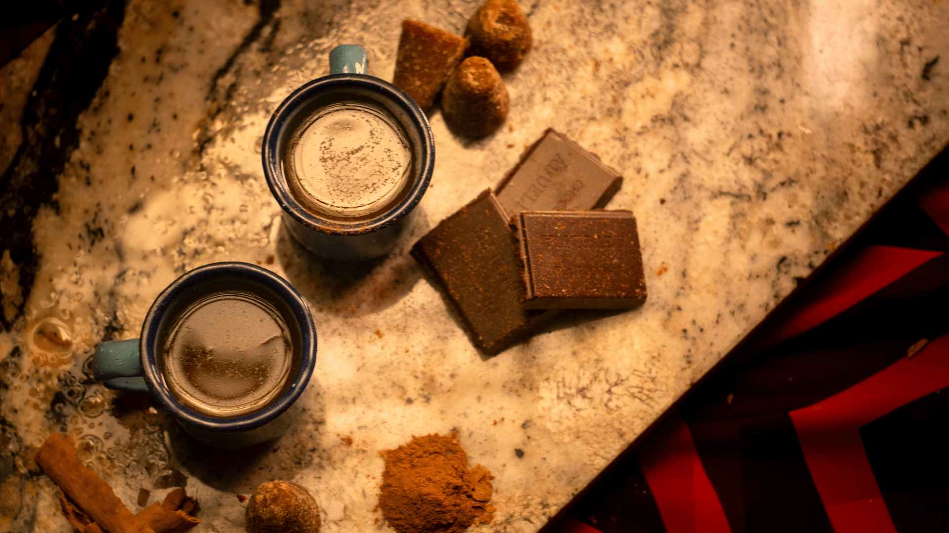 two mugs of hot coffee with chocolate pieces, cinnamon, and sugar arranged on a rustic surface