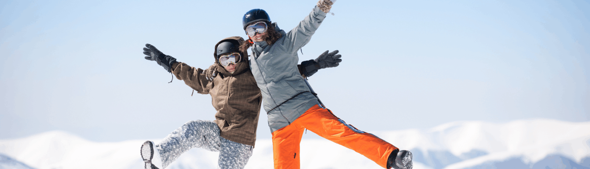 two people posing playfully on snowy mountain in ski gear with bright blue sky and snow covered peaks