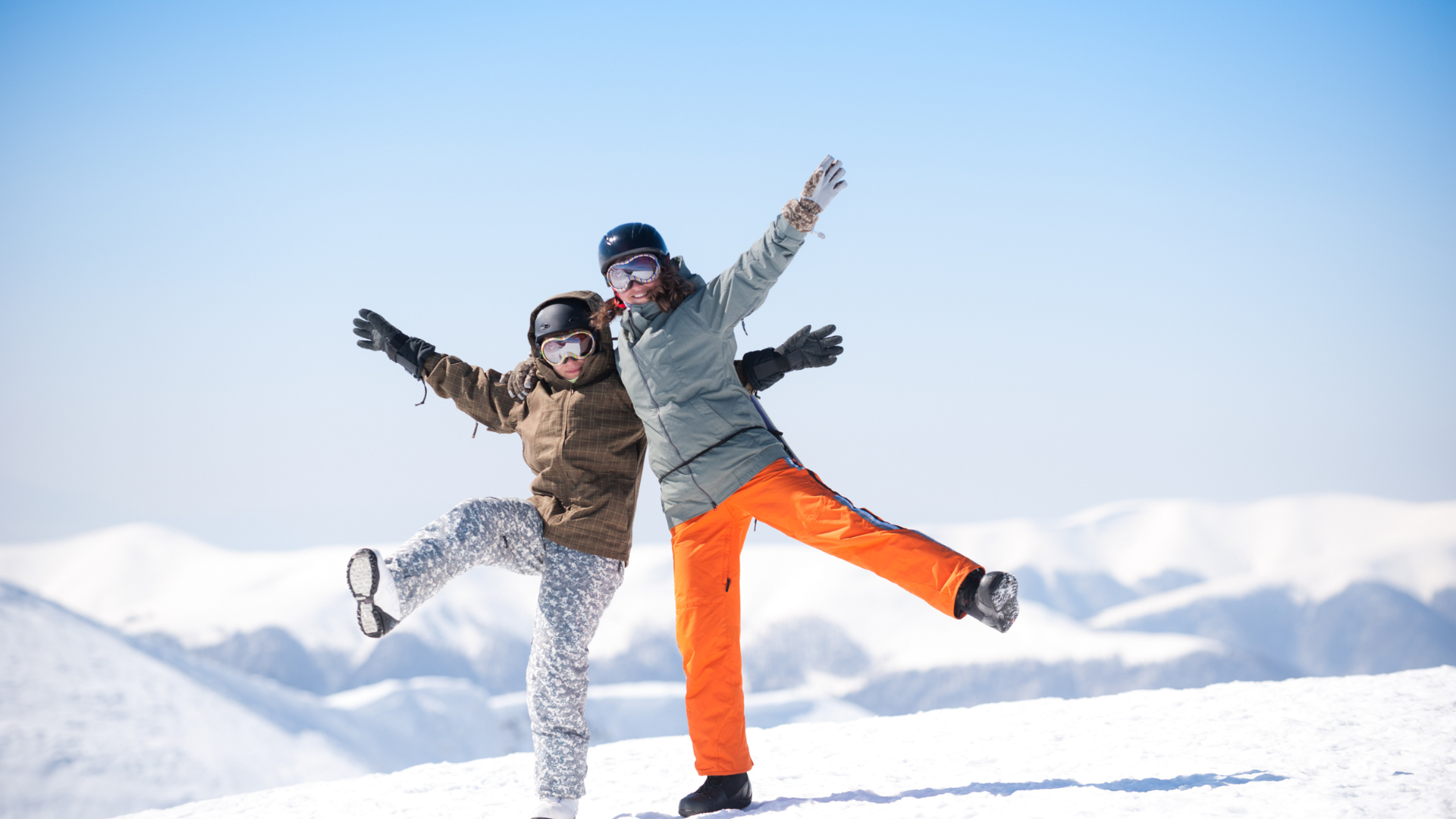 two people posing playfully on snowy mountain in ski gear with bright blue sky and snow covered peaks