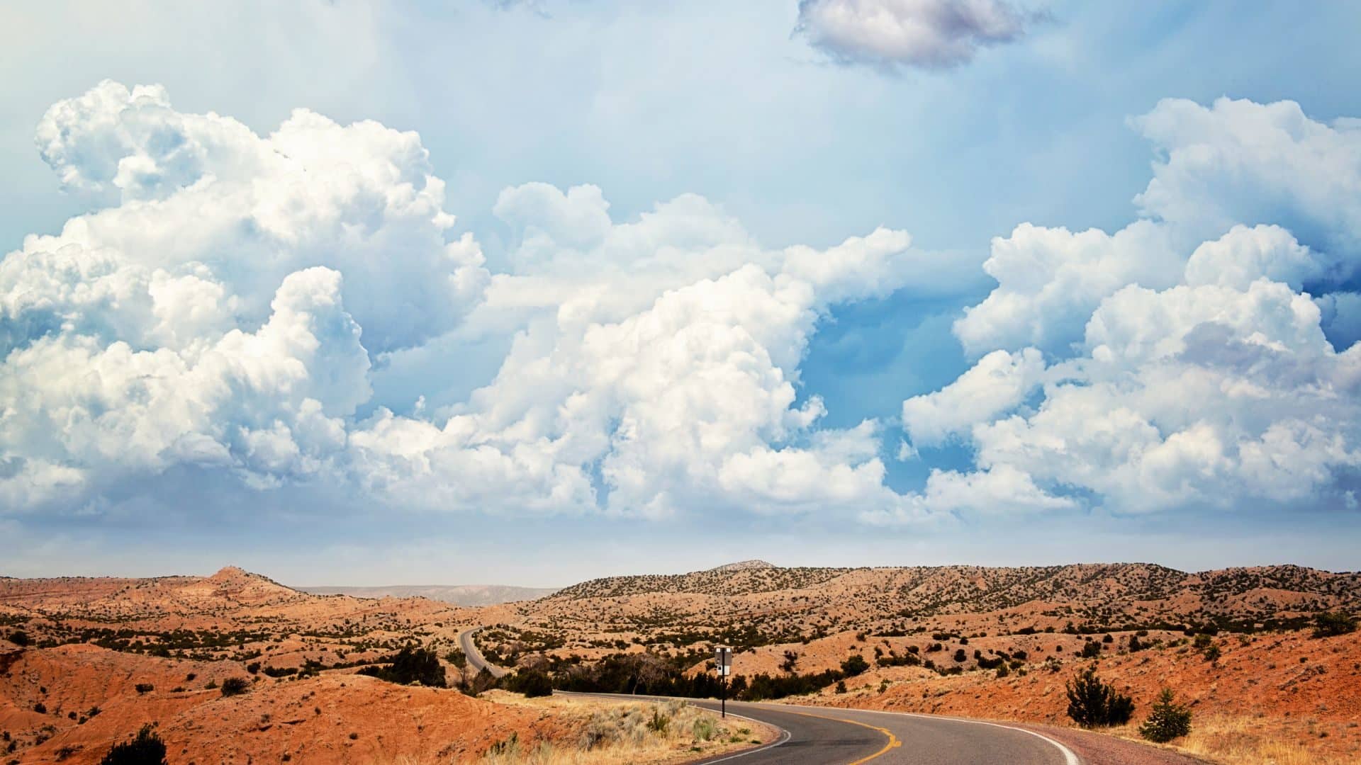 Road winding through brilliant orange topography against a backdrop of blue skies with large white puffy clouds.