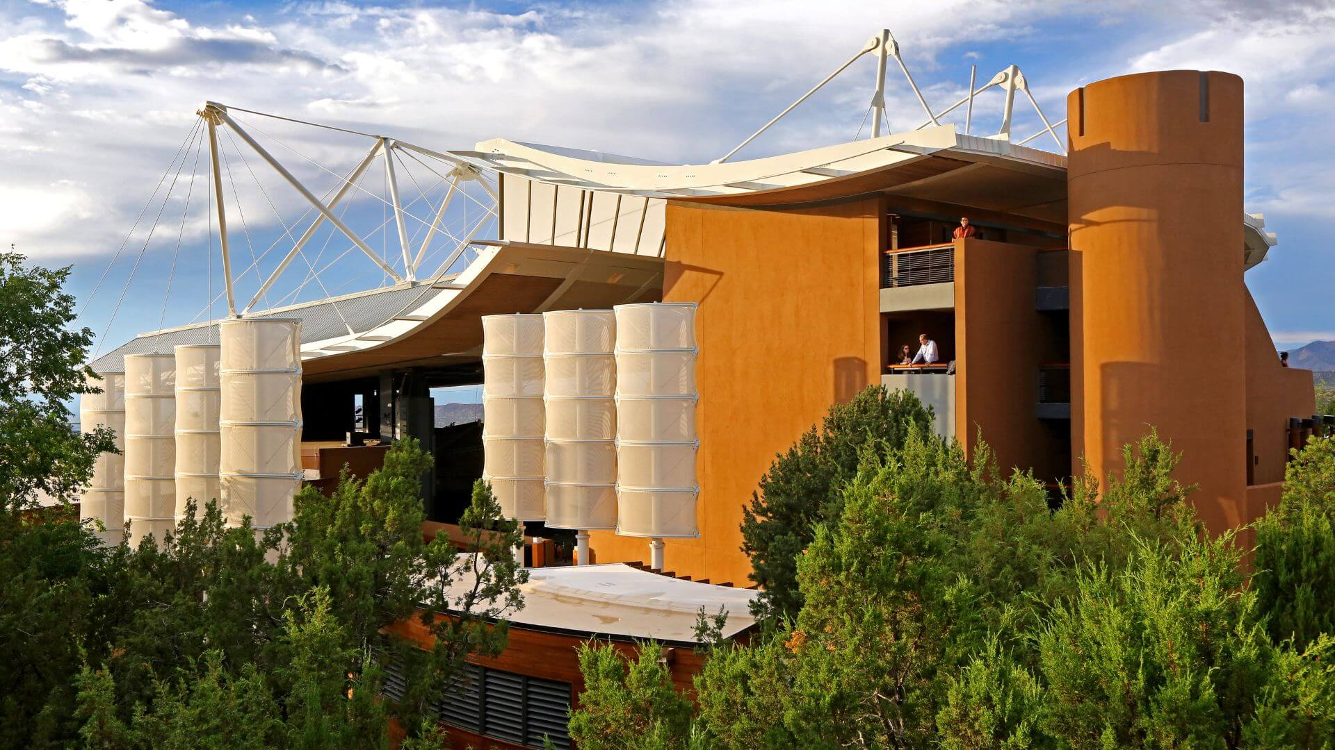 Santa Fe Opera House with modern open air design, curved roof structures, and white tensile elements, surrounded by trees under a partly cloudy sky