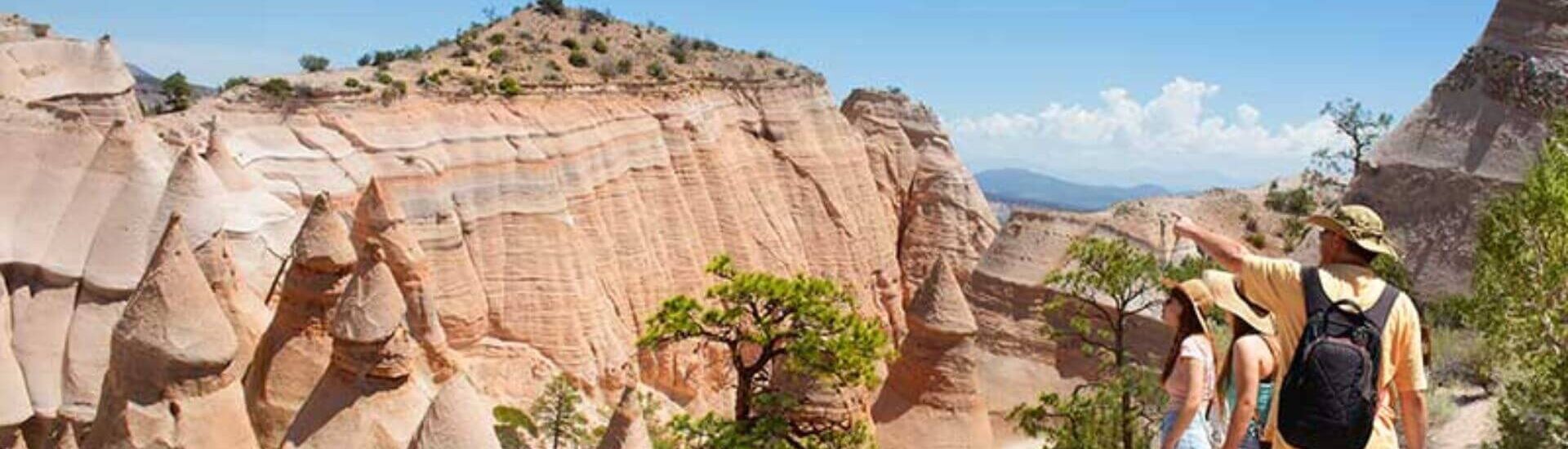 Hikers standing on a trail overlooking tent-shaped rock formations and layered canyon cliffs under a clear blue sky