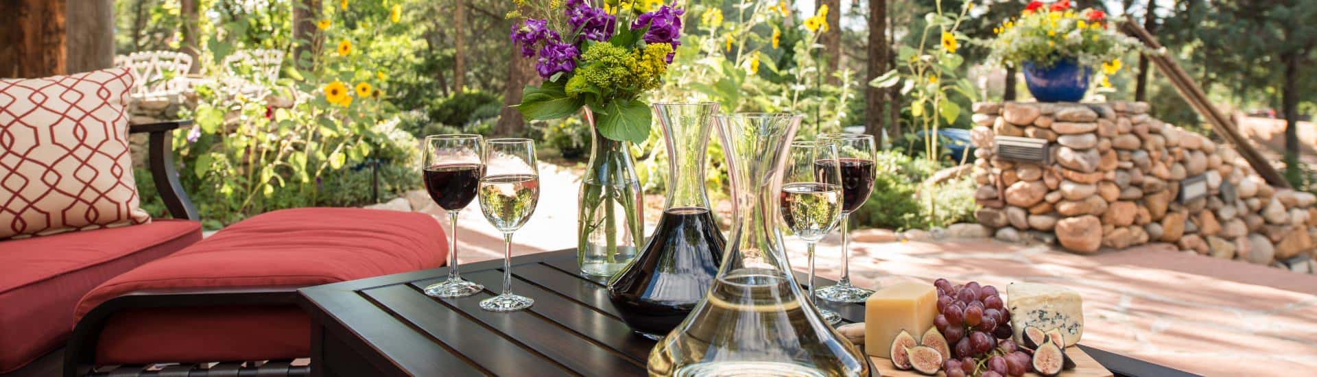 Dark wooden coffee table on stone patio with white and red wine decanters, glasses of poured white and red wine, and wooden tray with cheese and grapes